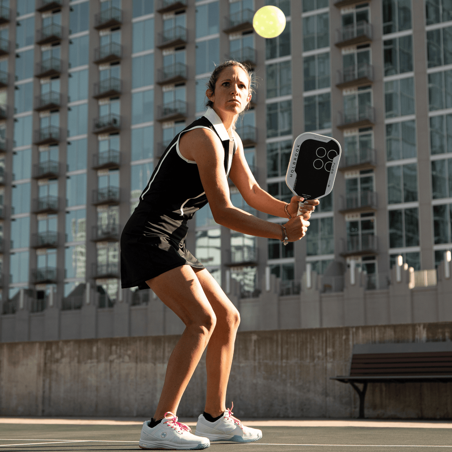 A woman in athletic clothing readies to hit a pickleball with the Holbrook Fuze Elongated 14mm Pickleball Paddle, featuring unibody carbon fiber construction, on an outdoor court with a city building in the background.