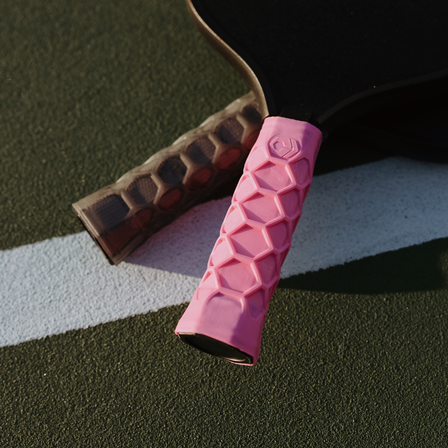 Two pickleball paddles on a court: one with a textured black grip, the other featuring Hesacore's XS Pink Pickleball Grip with a geometric pattern, lying on the dark green surface near a white line.