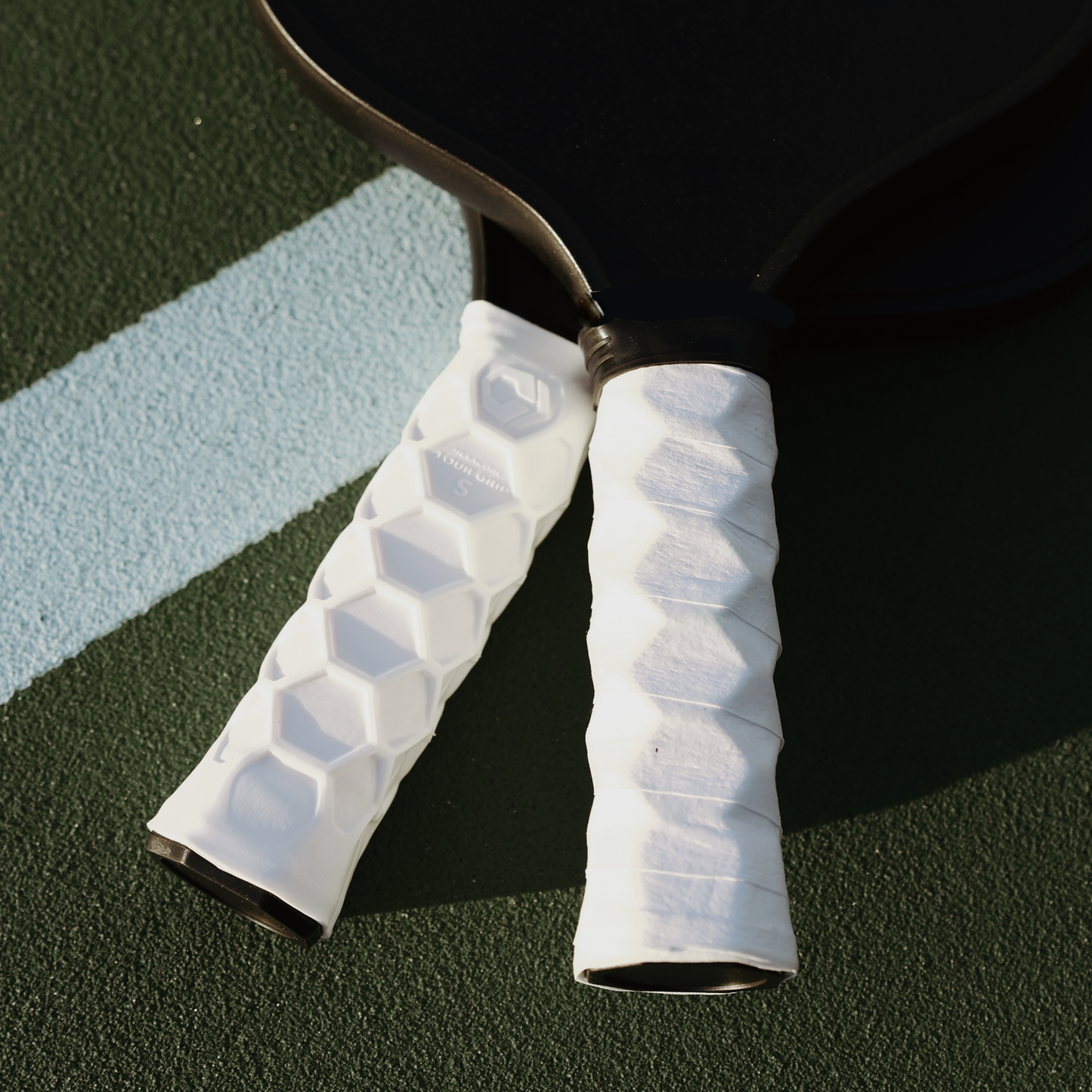 Two pickleball paddles featuring Hesacore Original White Pickleball Grips with textured surfaces rest on an outdoor court, partially positioned on a blue line.