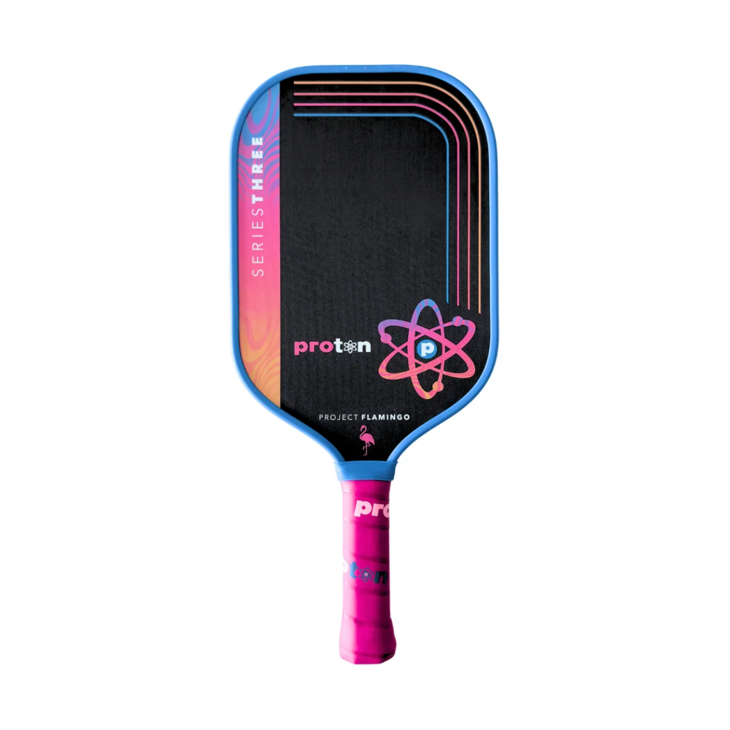 The Proton Project Flamingo Elongated 15mm Series Three Pickleball Paddle by Proton features a black carbon fiber face with blue edges, pink accents, a stylized atom graphic, pink grip tape, and high-density foam for better play.