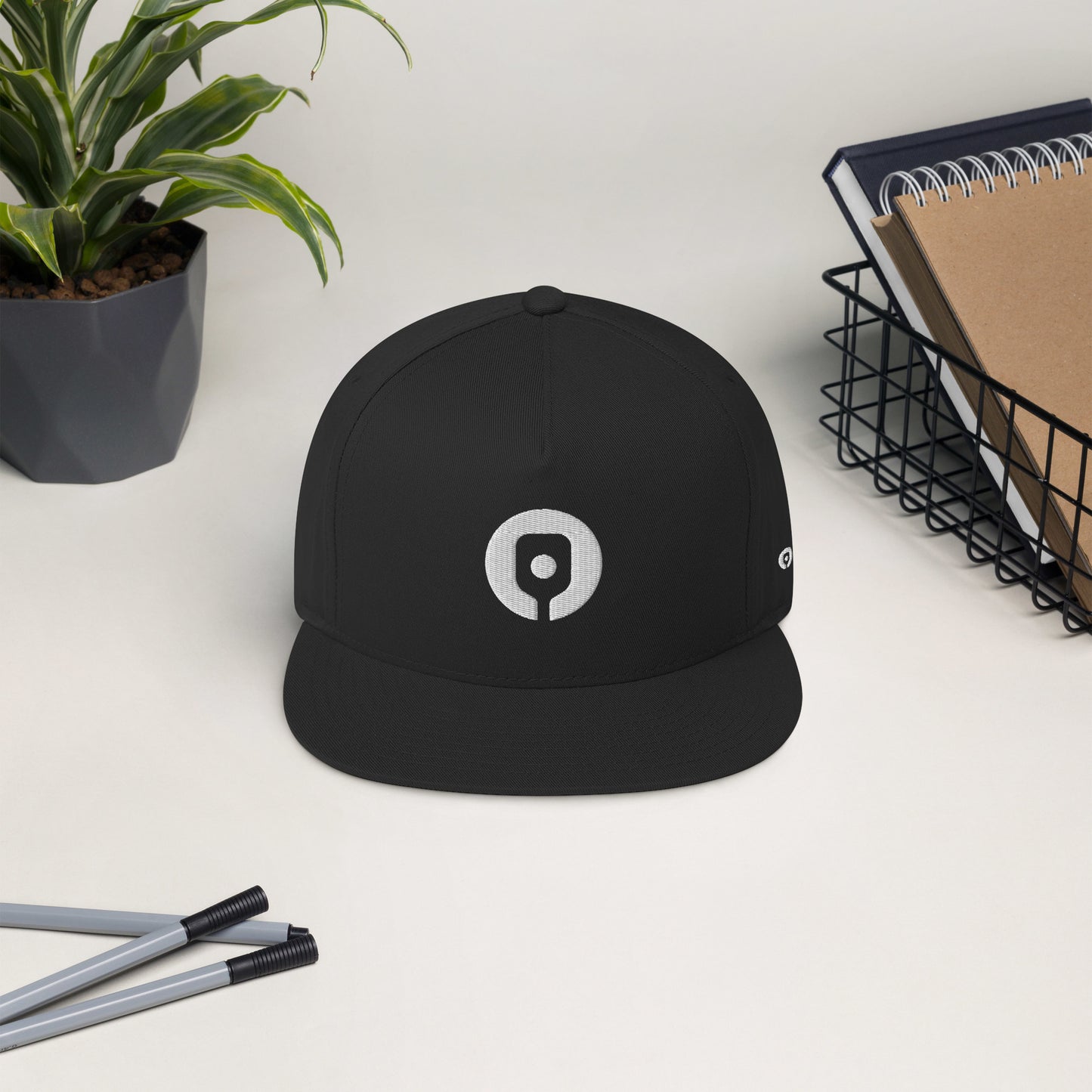 A Pickleballist Signature Logo Flat Brim Pickleball Hat in black sits on a desk alongside a potted plant, two pens, and a wire basket containing notebooks.