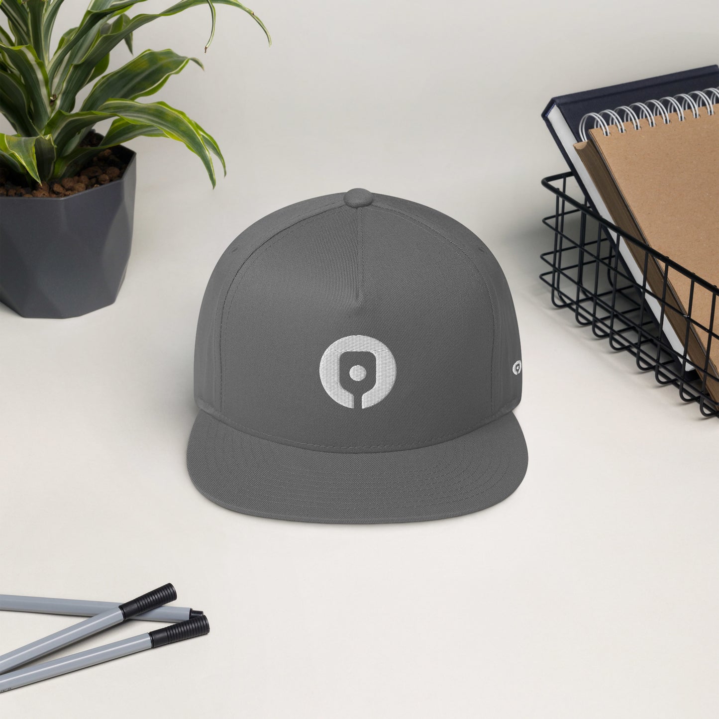 The Pickleballist Signature Logo Flat Brim Pickleball Hat, featuring a high-profile fit and a white circular logo, is positioned on a desk near a potted plant, pens, and a wire basket containing notebooks. Its modern gray flat-bill design enhances its contemporary appeal.
