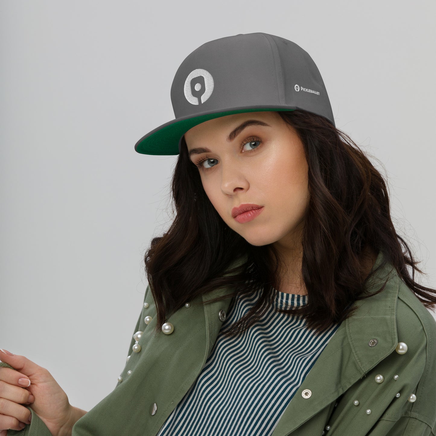 A person with long dark hair wearing the Pickleballist Signature Logo Flat Brim Pickleball Hat by Pickleballist, a green jacket adorned with pearls, and a striped shirt, posing against a plain background.