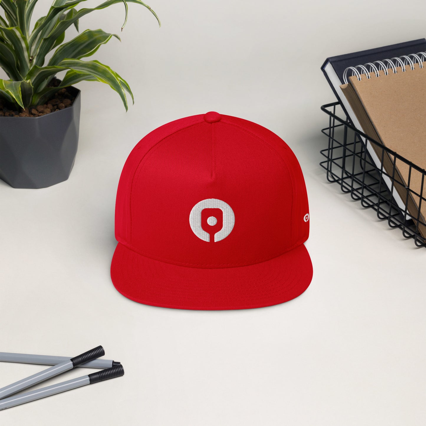 A Pickleballist Signature Logo Flat Brim Pickleball Hat featuring a white circular logo on the front is placed on a white surface. Nearby are two pens, a plant in a pot, and a wire basket containing notebooks. This high-profile fit from Pickleballist includes a snapback closure for easy adjustment.