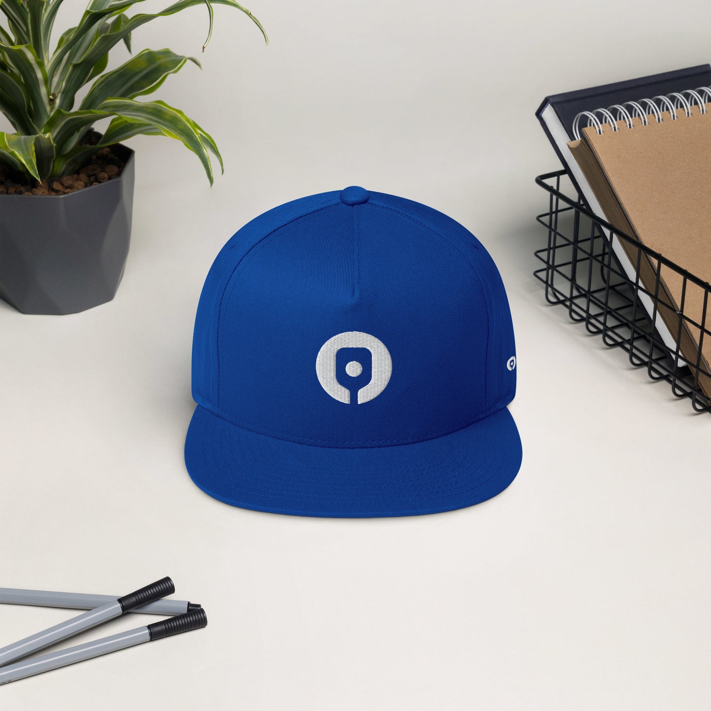 The Pickleballist Signature Logo Flat Brim Pickleball Hat in blue, featuring a white circular logo, sits on a white desk alongside a potted plant, pens, and a wire basket holding notebooks. This high-profile fit hat from Pickleballist also includes a snapback closure for customizable comfort.