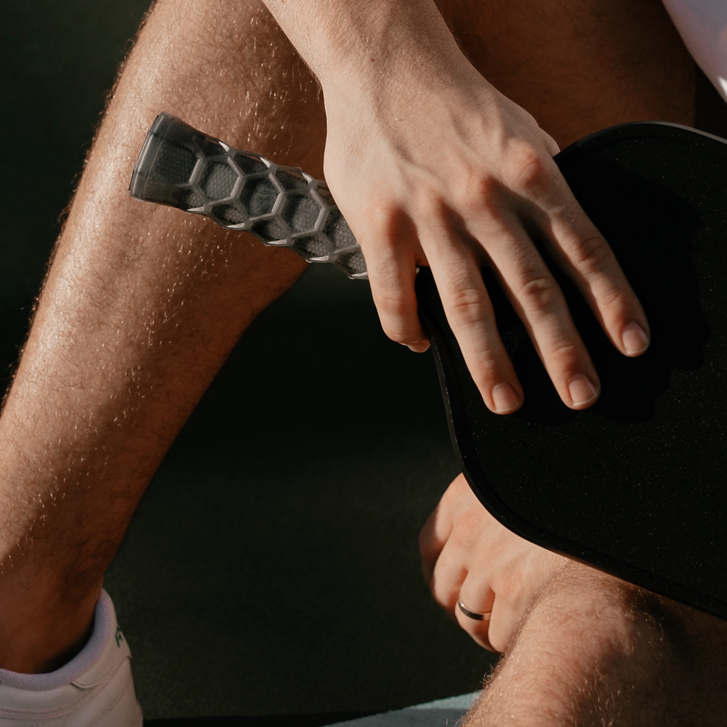 A person sits cross-legged, holding a pickleball paddle with a Hesacore Carbon (Charcoal) Pickleball Grip, their hand resting on their knee.
