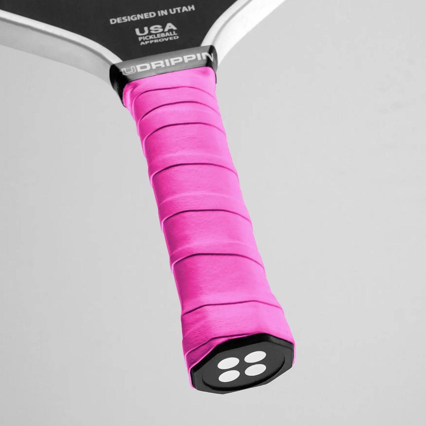 Close-up of a black pickleball paddle handle wrapped in UDRIPPIN Comfort Tour Overgrips, featuring a bright pink color, against a gray background. Text on the paddle reads "Designed in Utah, USA Pickleball Approved.