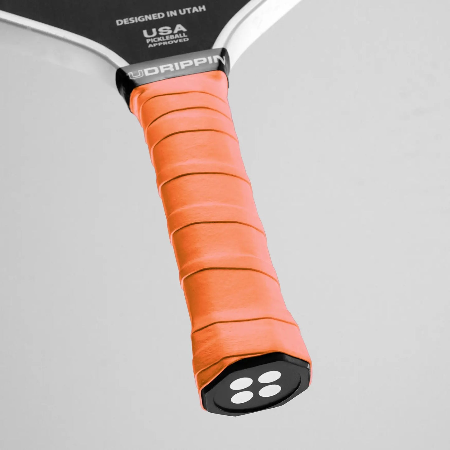 Close-up of a pickleball paddle handle featuring the UDRIPPIN Comfort Tour Overgrips in orange, with a label indicating "Designed in Utah, USA, Pickleball Approved.