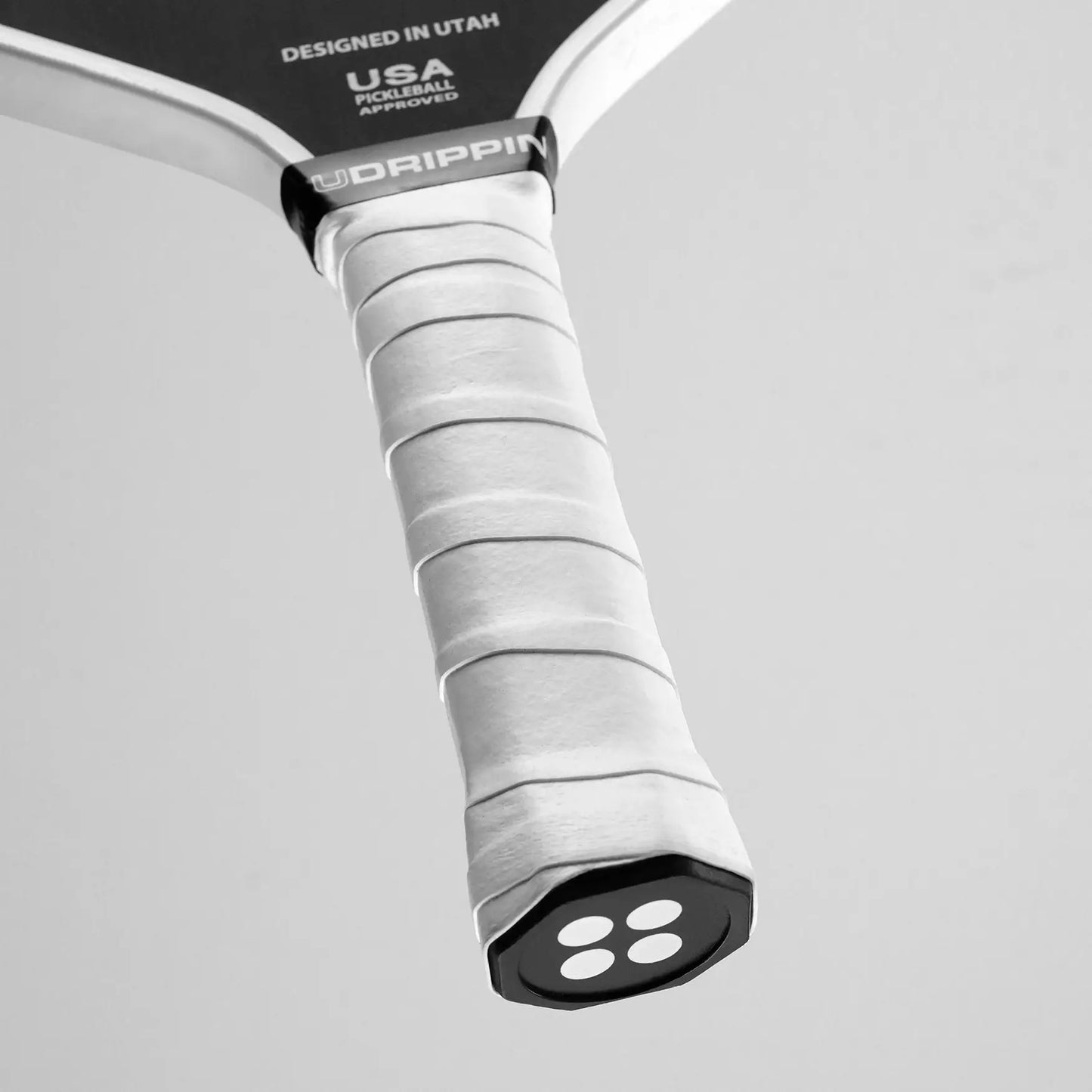 Close-up of a pickleball paddle handle featuring the UDRIPPIN Pro Tour Extra Tacky Overgrips in white. The base of the handle displays an embossed logo with circular designs.