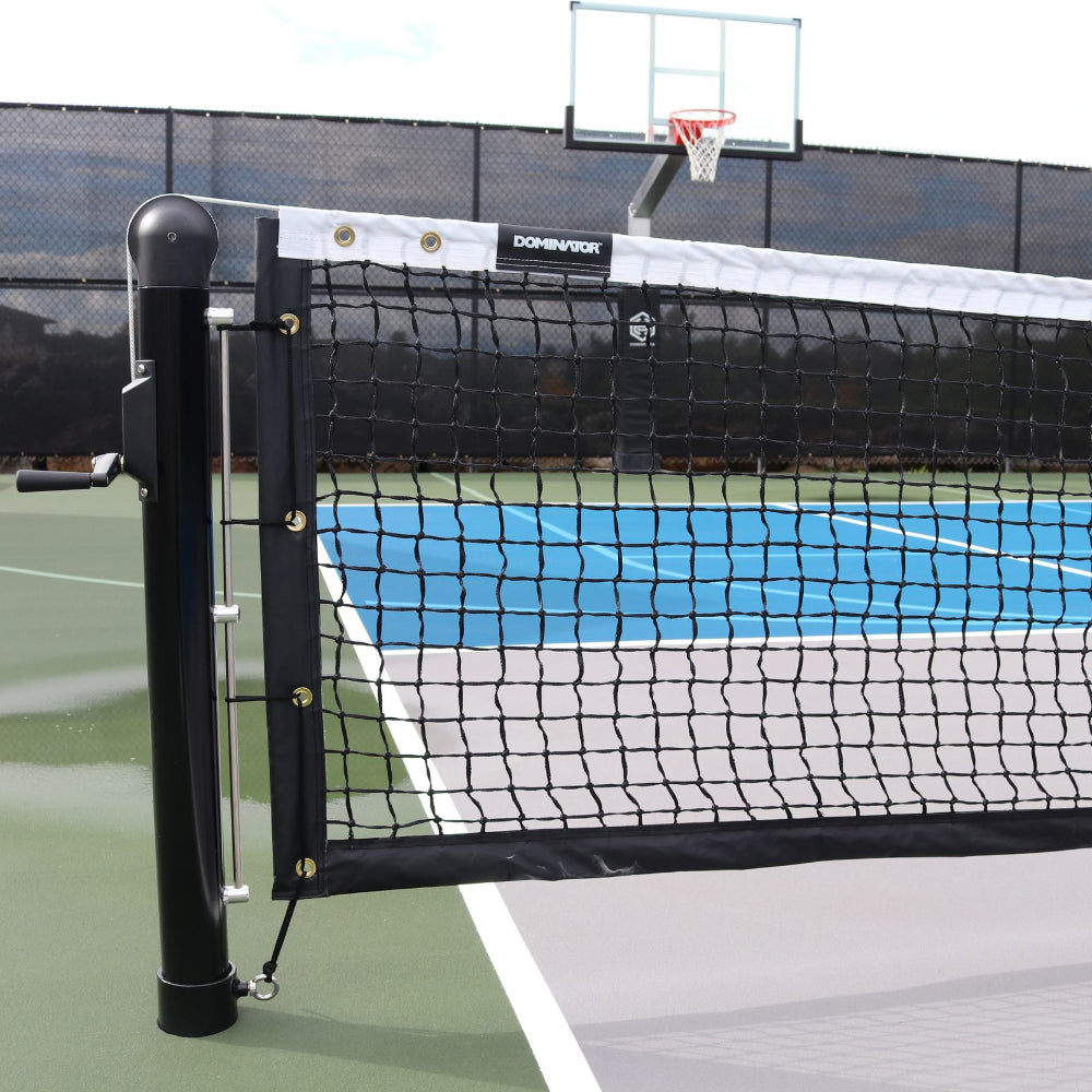 Close-up of Dominator In-Ground Pickleball Posts & Nets System with a crank handle on a multi-sport court featuring a basketball hoop in the background.