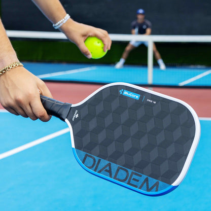 Close-up: Someone holds the Diadem Edge BluCore Pro 14mm Pickleball Paddle and a yellow ball on a blue court, with another player in the background.