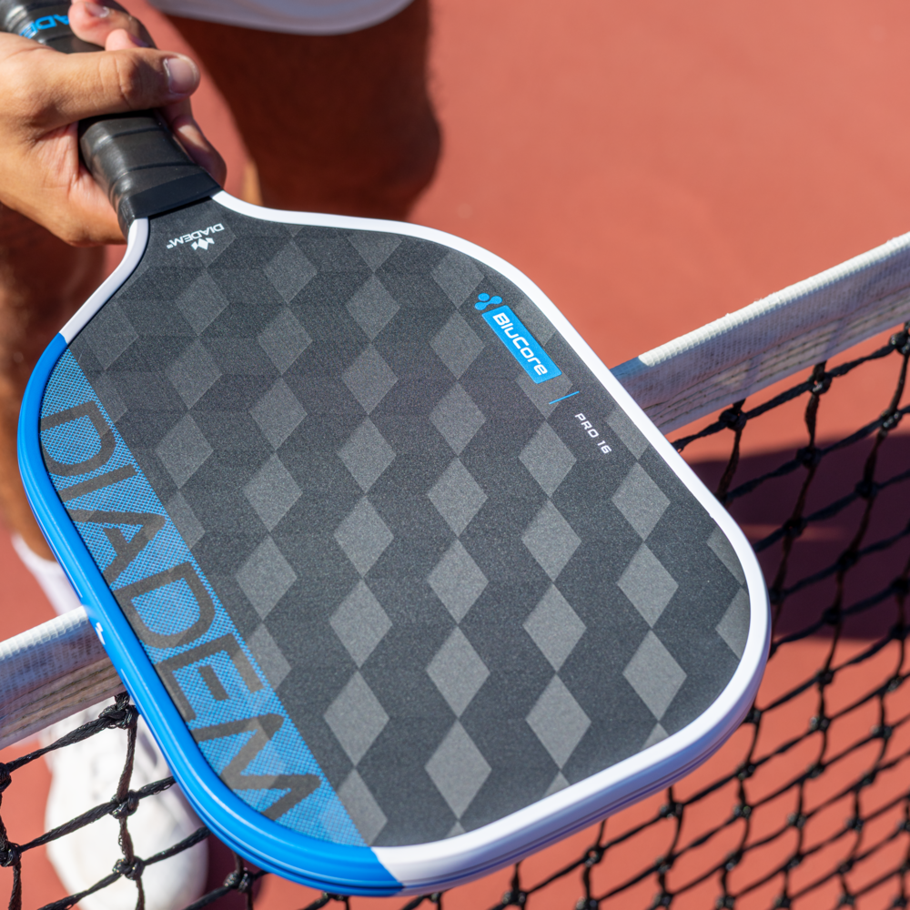 A person holds a Diadem Edge BluCore Pro 14mm Pickleball Paddle over a net on an outdoor court, highlighting its sleek design and high performance.