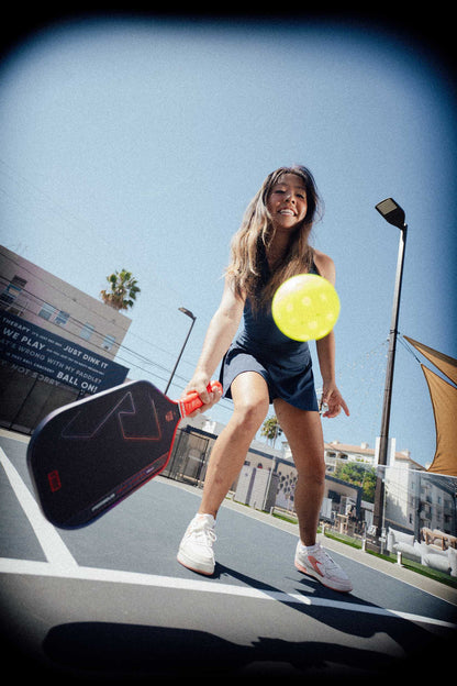 A woman on an outdoor pickleball court swings a JOOLA Heat Vision Perseus 16mm Pickleball Paddle at a yellow ball, playing a forehand shot under a clear blue sky.