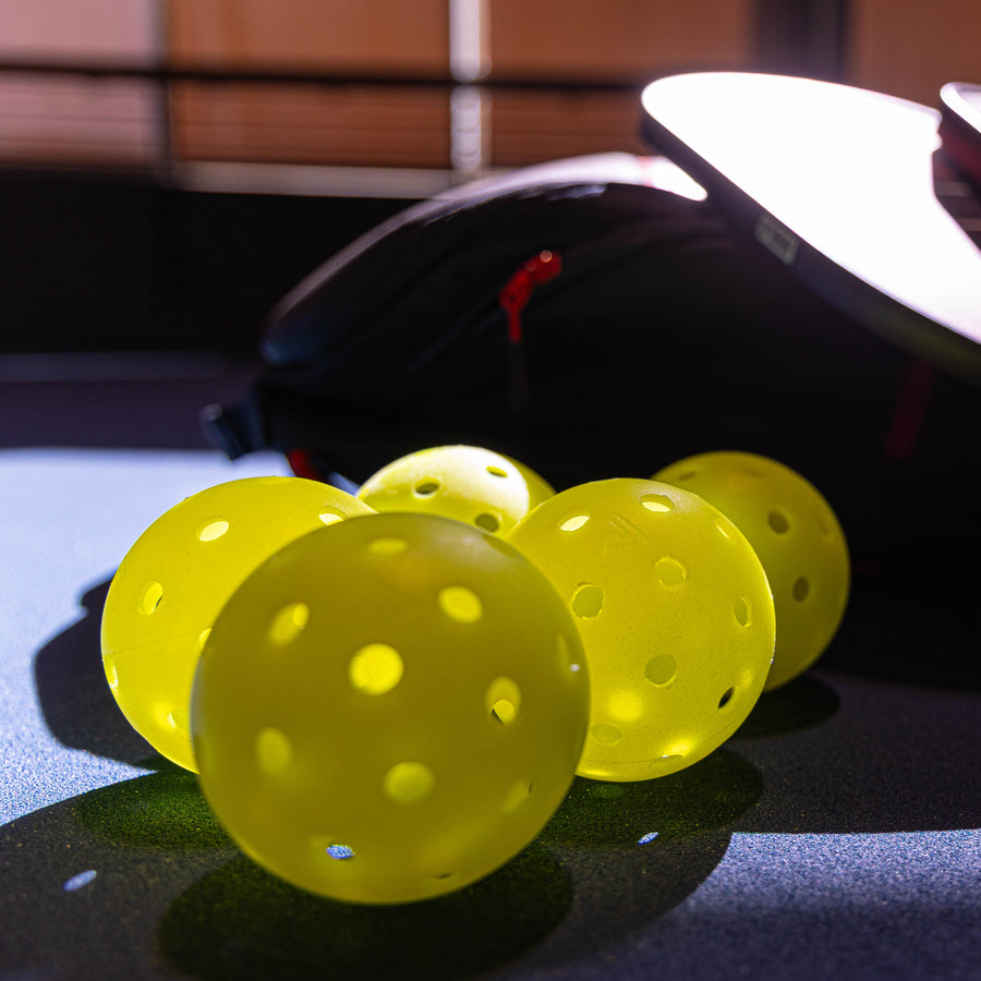 Four Pickleballist JOOLA HC-40 Shock Yellow pickleballs rest on a blue surface, with a black sports bag and paddle behind—ideal for players needing durable tournament balls.