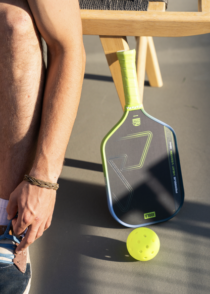 A person sits next to a Pickleballist JOOLA HC-40 Pickleball paddle and Shock Yellow pickleballs on the ground, sunlight casting gentle shadows.