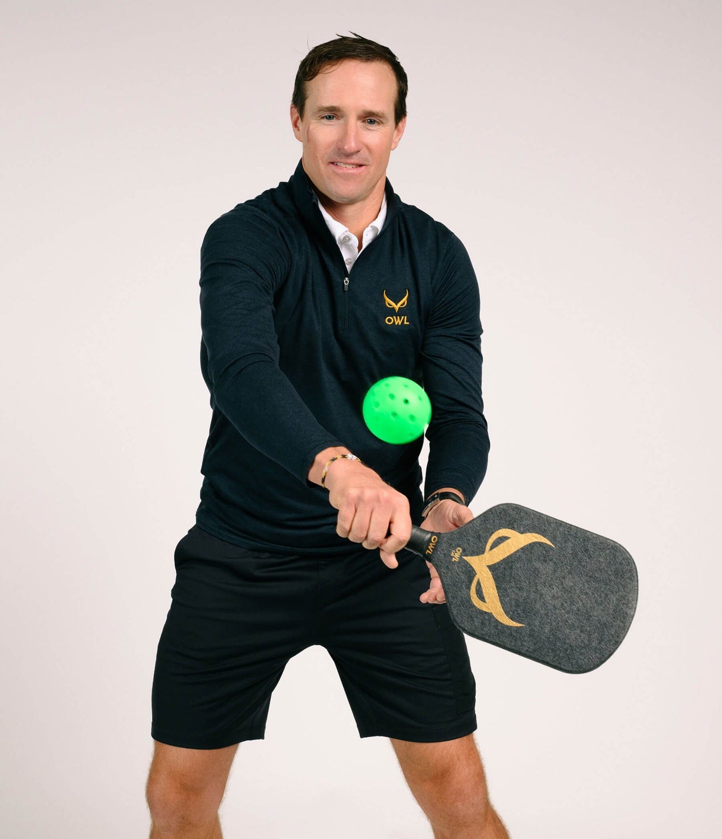 A man in a dark athletic outfit strikes a green pickleball with the OWL CXE - Drew Brees Limited Edition Quiet Pickleball Paddle by OWL, standing against a plain light background.