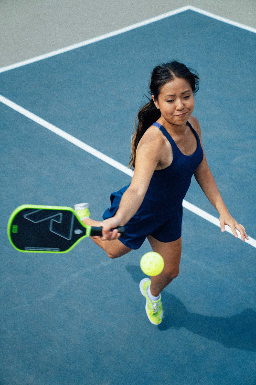 A woman in athletic wear uses the JOOLA Double Vision Hyperion 16mm Pickleball Paddle to hit a yellow pickleball on an outdoor court.