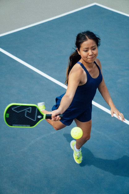 A woman in athletic wear uses the JOOLA Double Vision Hyperion 16mm Pickleball Paddle to hit a yellow pickleball on an outdoor court.