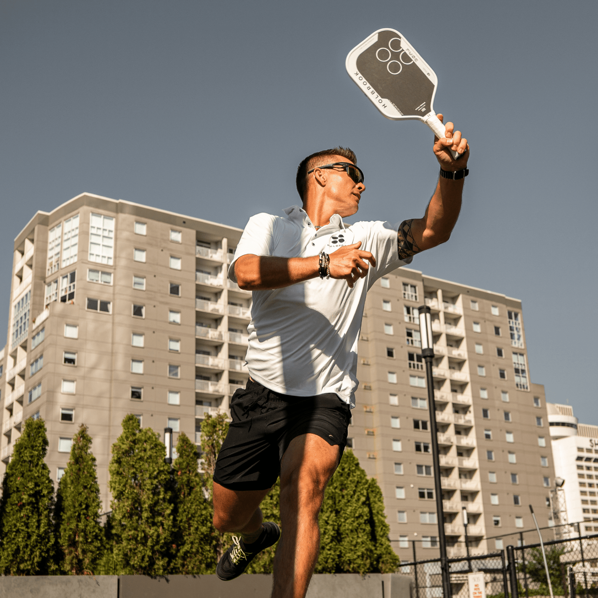 A man in athletic wear plays pickleball outdoors, swinging his Holbrook Fuze Elongated 14mm Pickleball Paddle as buildings and trees frame the action.
