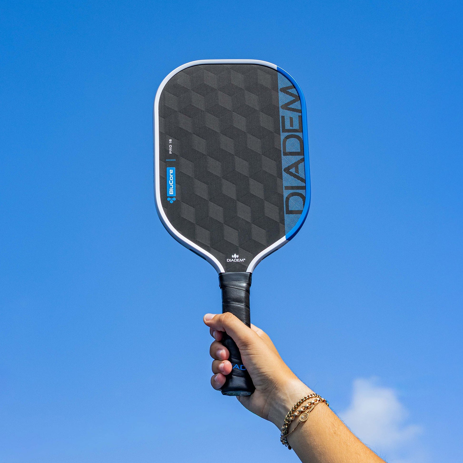 A hand holds up the Diadem Edge BluCore Pro 14mm Pickleball Paddle against a clear blue sky, highlighting the sleek design of this premium paddle from Diadem.