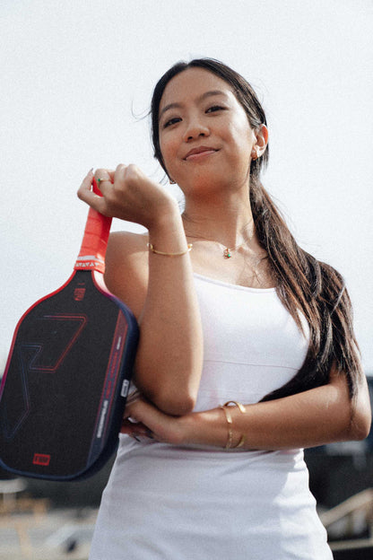 A woman in a white tank top smiles confidently at the camera, holding a JOOLA Heat Vision Scorpeus 16mm Pickleball Paddle by JOOLA over her shoulder.