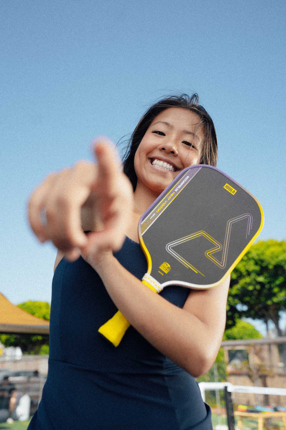 A smiling woman holds a JOOLA Heat Vision Scorpeus 16mm Pickleball Paddle and points toward the camera outside on a sunny day.