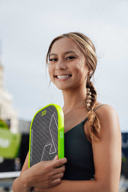 A young woman with a braided hairstyle smiles while holding the JOOLA Double Vision Scorpeus 16mm Pickleball Paddle outdoors in casual athletic wear—ideal for advanced players.