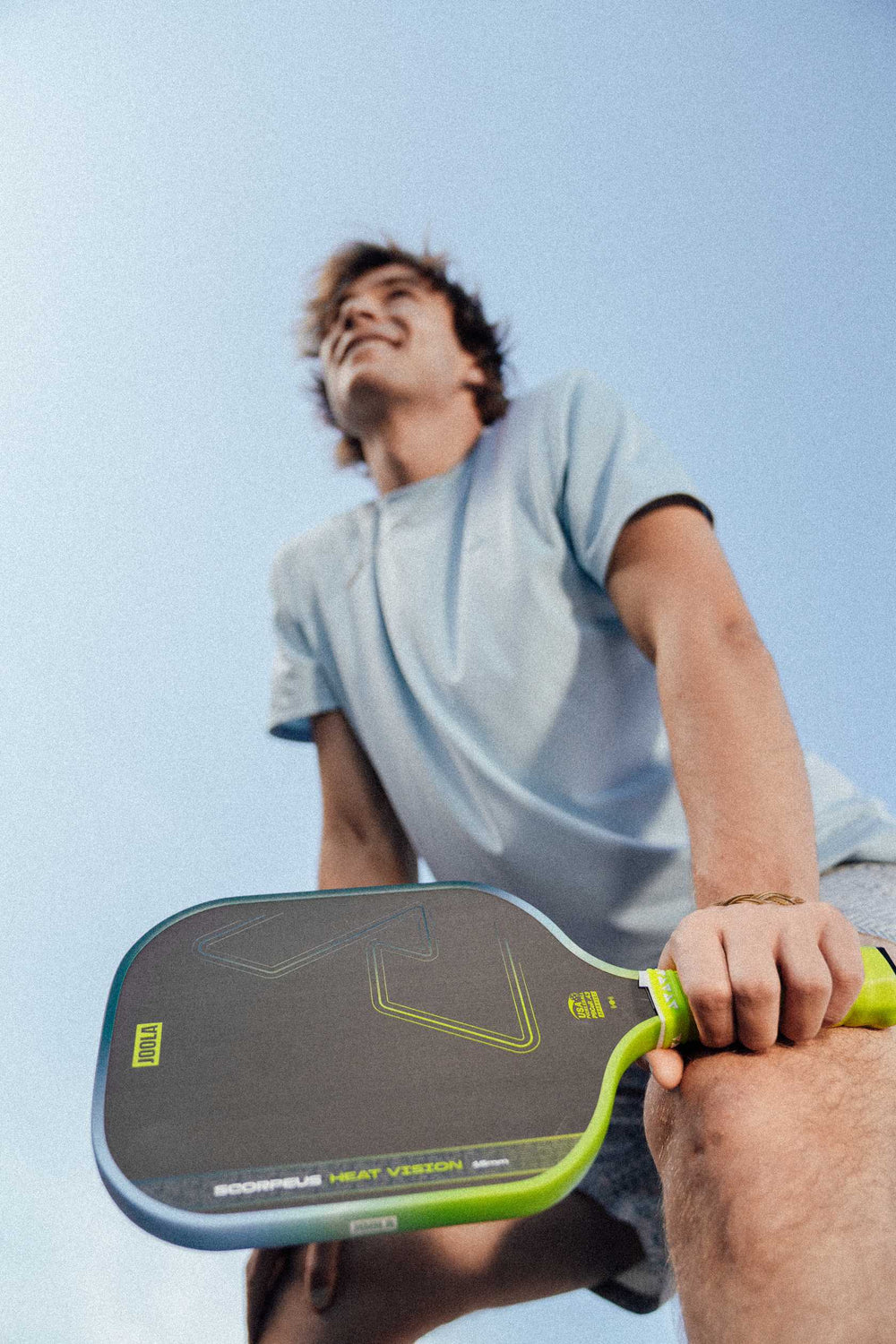A person in a light blue shirt holds a JOOLA Heat Vision Scorpeus 16mm Pickleball Paddle, viewed from a low angle against a blue sky.