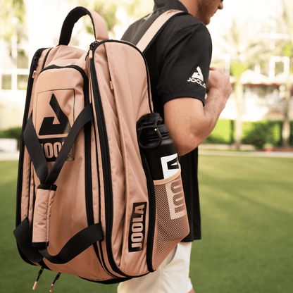 A person carries a beige Pickleballist 2026 Tour Elite Pickleball Bag with a black water bottle in the side pocket, walking on green grass and wearing a black JOOLA shirt.
