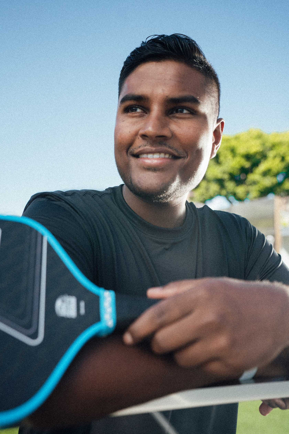 A man in a dark shirt smiles while leaning on a table, holding the JOOLA Double Vision Hyperion 16mm Pickleball Paddle by JOOLA, with trees and a clear sky in the background.