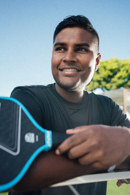 A man in a dark shirt smiles while leaning on a table, holding the JOOLA Double Vision Hyperion 16mm Pickleball Paddle by JOOLA, with trees and a clear sky in the background.