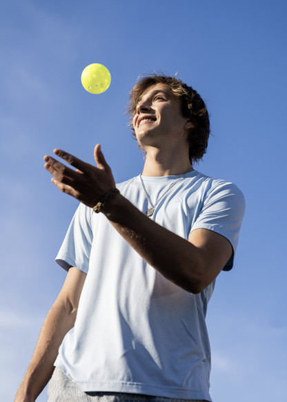 A young person in a light blue shirt tosses a Pickleballist JOOLA HC-40 Pickleball in Shock Yellow into the air while looking up, with a blue sky in the background.