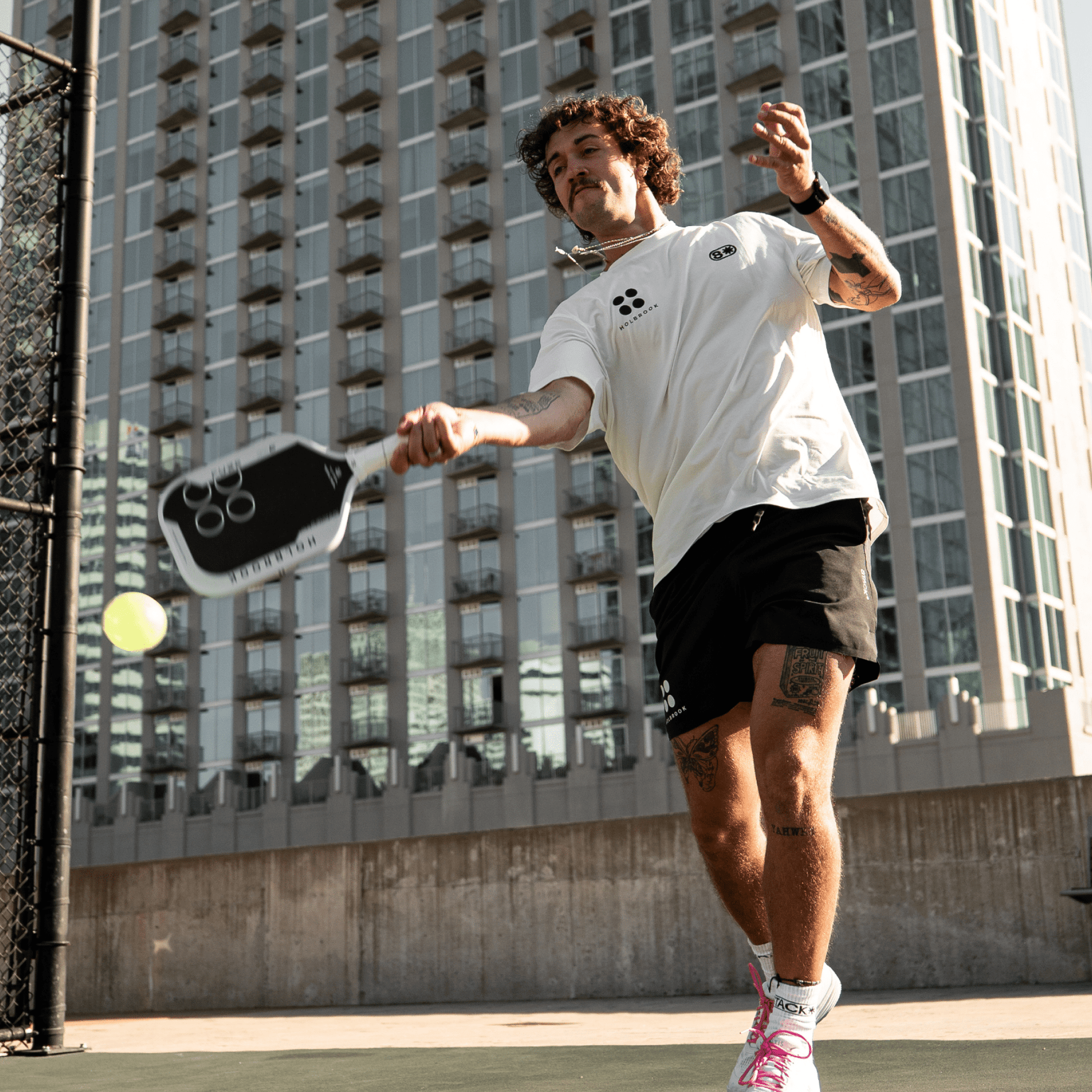 A man in athletic wear plays pickleball on an outdoor court in front of a tall apartment building, about to hit the ball with his Holbrook Fuze Hybrid 16mm Pickleball Paddle.