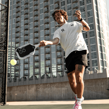 A man in athletic wear plays pickleball on an outdoor court in front of a tall apartment building, about to hit the ball with his Holbrook Fuze Hybrid 16mm Pickleball Paddle.