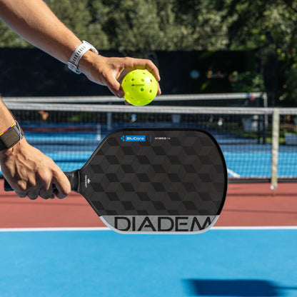 A person holds a yellow pickleball and a Diadem Edge BluCore Hybrid 16mm Pickleball Paddle by Diadem on an outdoor court with a net in the background, highlighting this vibrant paddle in action.