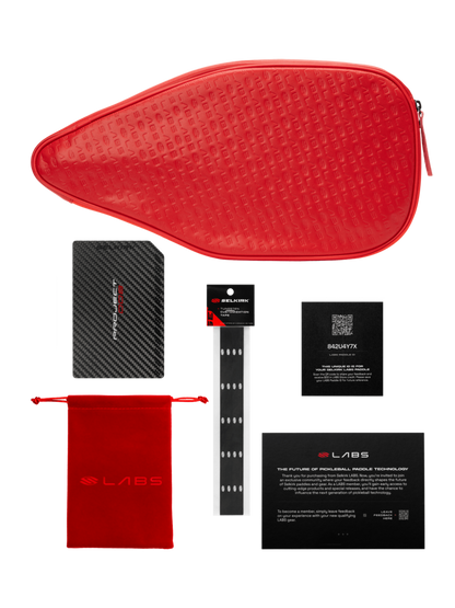 A red zippered pouch, black card, small red pouch, black wristband, black box, and a text card are artfully arranged with the Selkirk Labs Project 008 Invikta Elongated 13mm Pickleball Paddle by Selkirk on a white background.