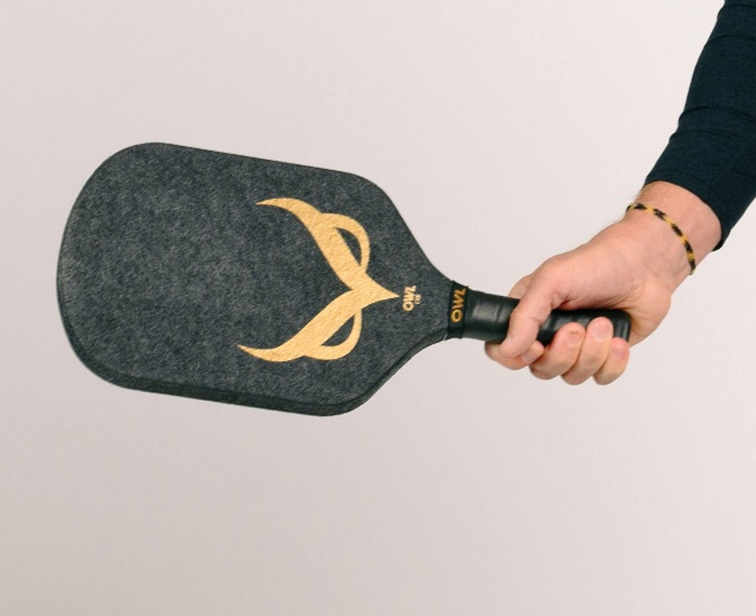 A person holding the OWL CXE - Drew Brees Limited Edition Quiet Pickleball Paddle by OWL, featuring a gold owl face design, black grip, and crafted from high-performance Propylux Acoustene composite.