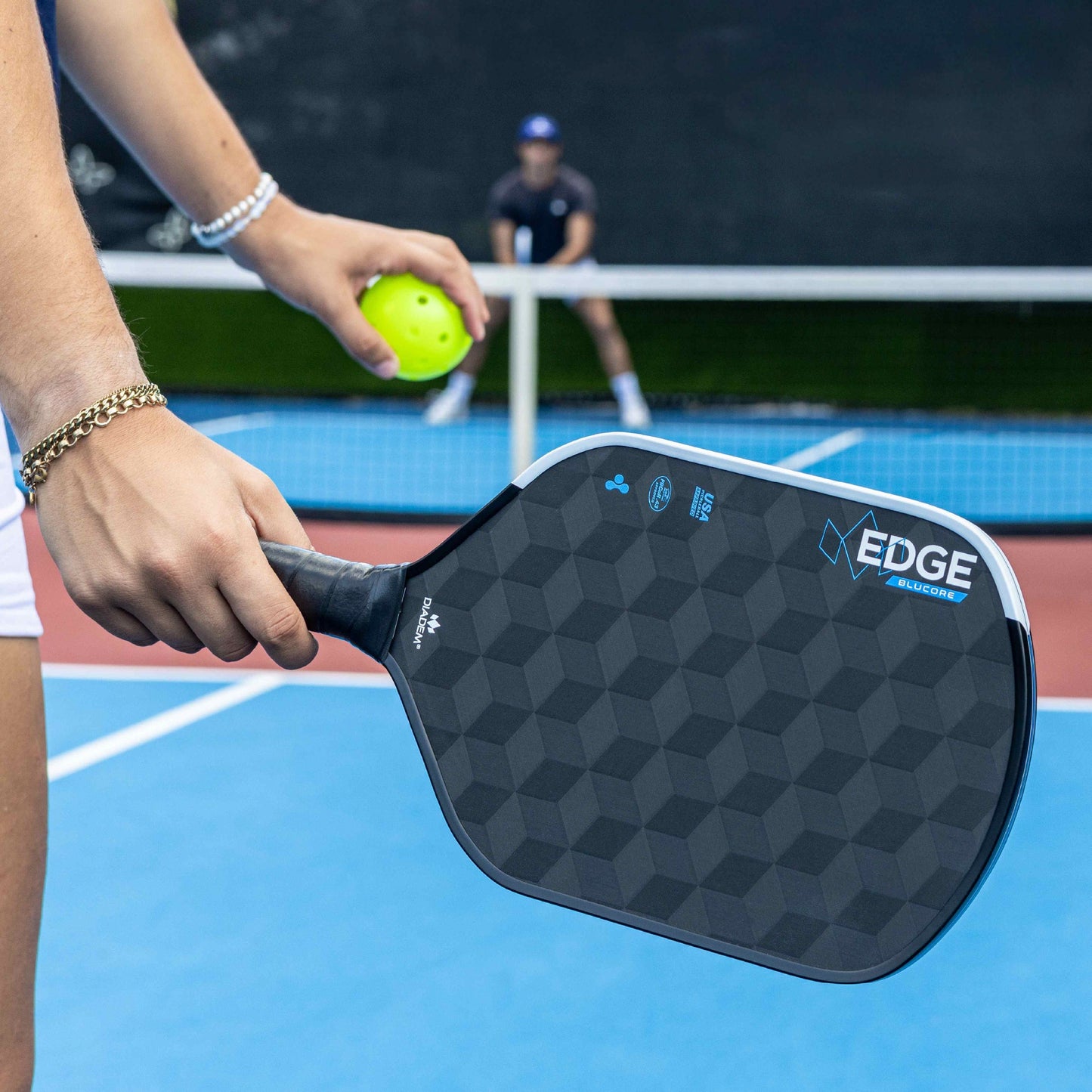 Close-up of the Diadem Edge BluCore Hybrid 16mm Pickleball Paddle by Diadem held with a ball on a blue court, while another player sits in the background.