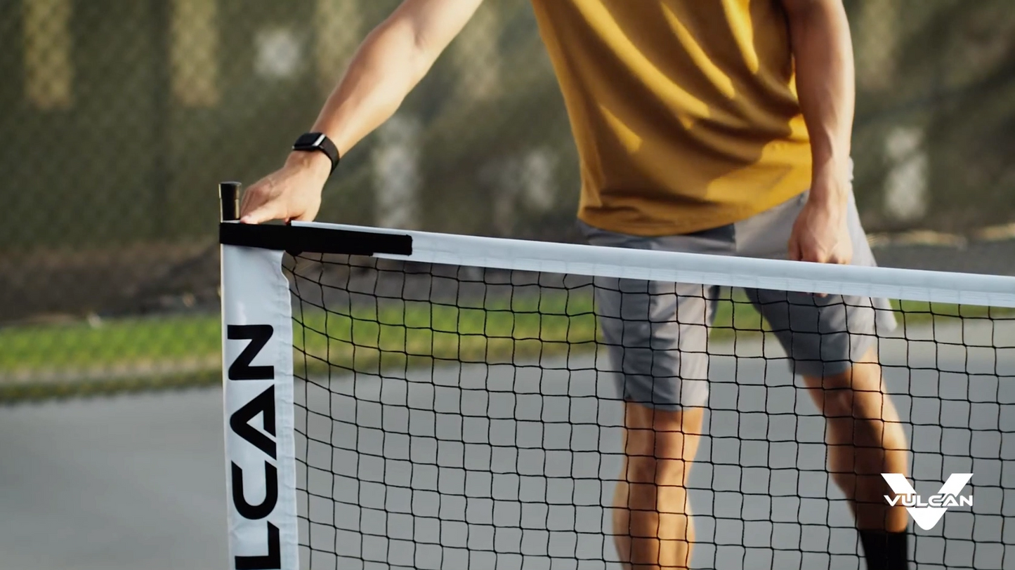 A person in a yellow shirt and gray shorts adjusting a Pickleballist Vulcan VNET on an outdoor court.