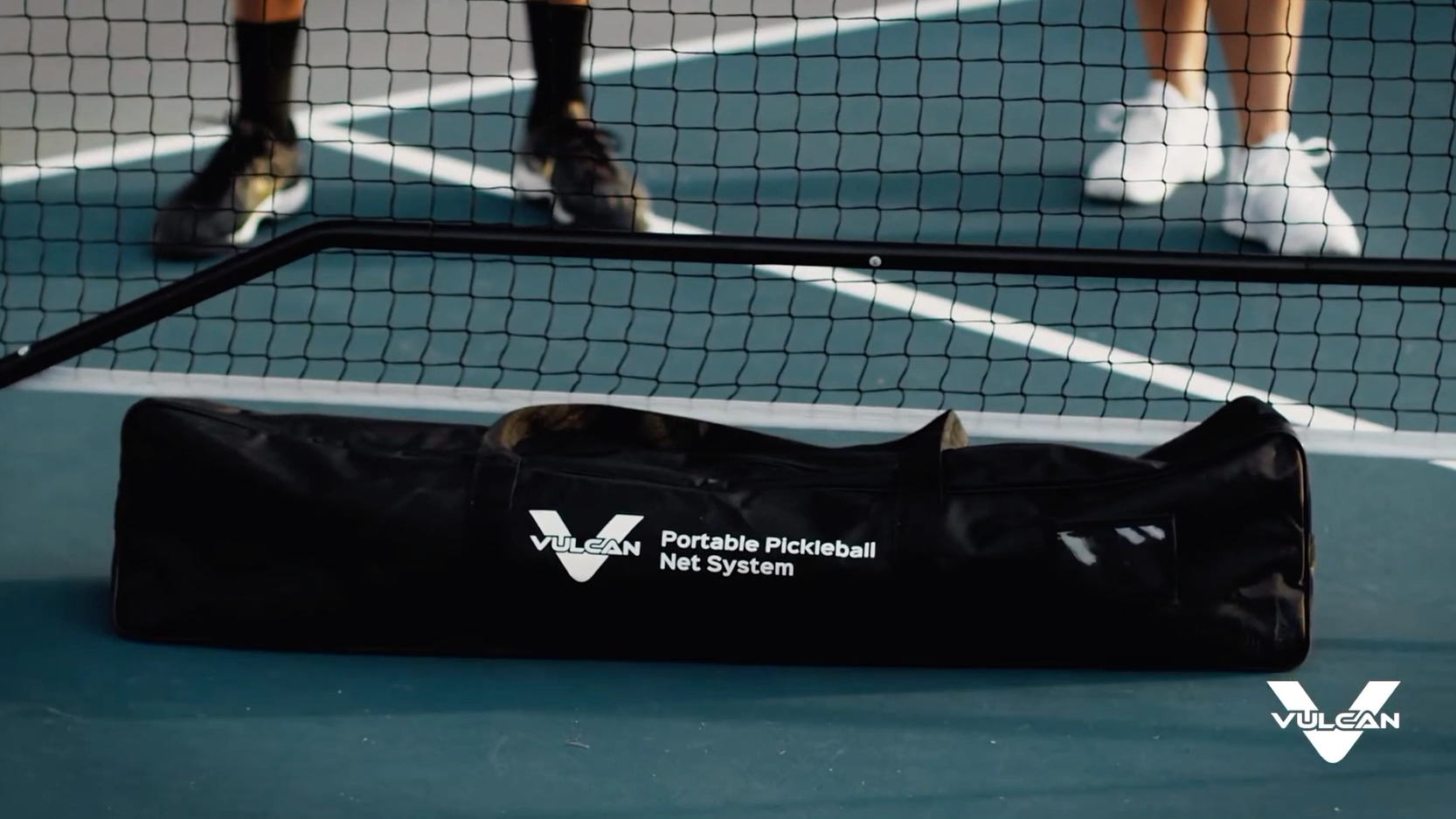 A Pickleballist Vulcan VNET Pickleball Net system bag, placed on a tennis court next to a net, with a person's legs visible in the background.