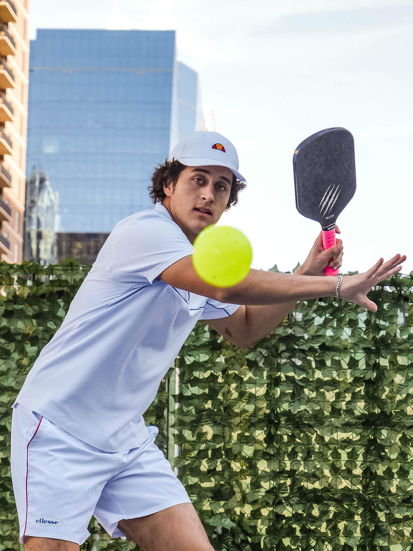 A man in a white sports outfit and cap gets ready to hit a pickleball with the OWL Talon X1 Quiet Pickleball Paddle, focusing on spin control, with a green hedge and tall buildings in the background.