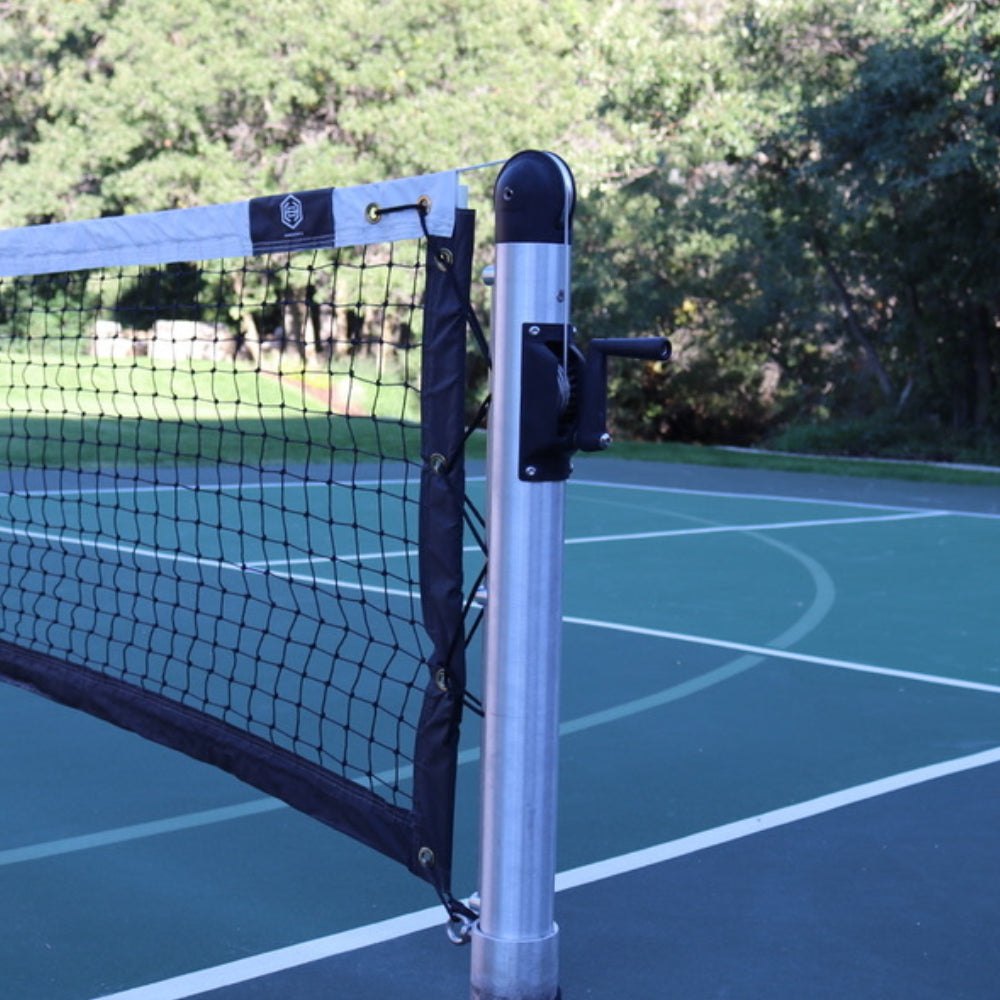 A close-up of a Dominator In-Ground Pickleball Posts & Nets System attached to a metal pole on an outdoor court with trees in the background.