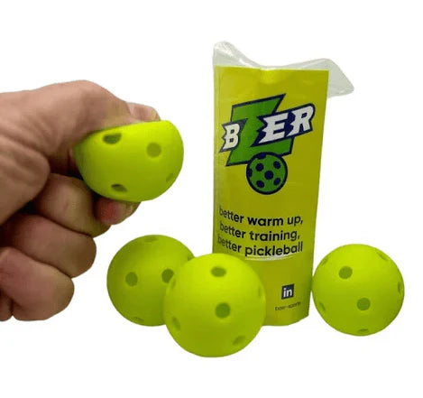 A hand squeezes a yellow BZER Mini Training Pickleball by Bzer Sports, with three balls and a canister labeled "BZer: better warm up, better training, better pickleball" in the background—ideal for boosting hand-eye coordination.
