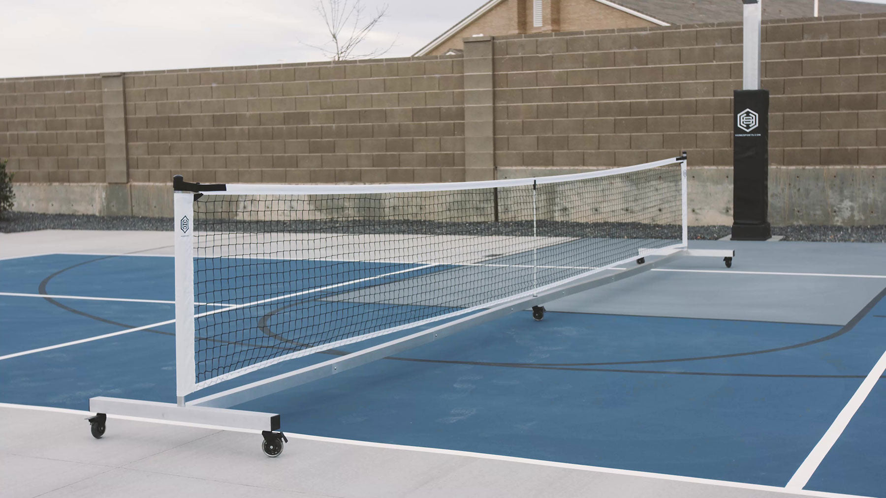 The Dominator Standard Portable Pickleball Net is set up on a blue and white outdoor court, with a tall brick wall in the background.